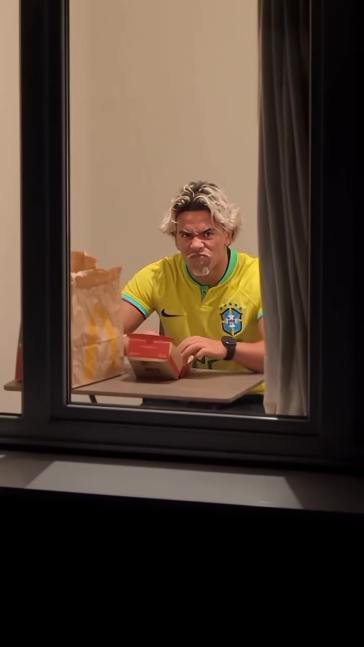 A person eating a burger in a lit window at night, caught by the camera